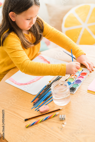 Little girl using watercolors to paint a rainbow wearing a yellow shirt in a bright and colorful home