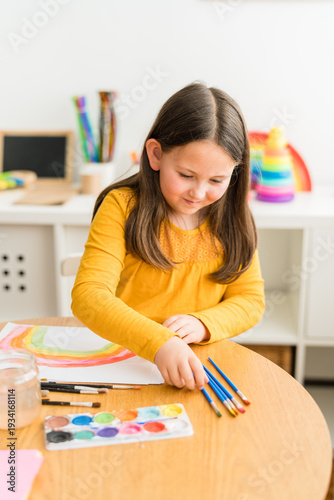 Little girl using watercolors to paint a rainbow wearing a yellow shirt in a bright and colorful home