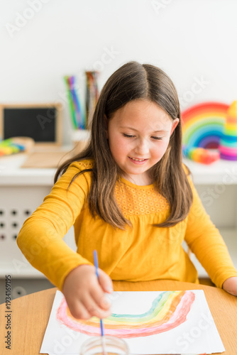 Little girl using watercolors to paint a rainbow wearing a yellow shirt in a bright and colorful home