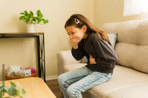 Little girl sitting on a sofa, clutching her stomach and covering her mouth, feeling unwell with symptoms of nausea and illness