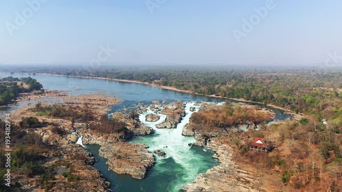 Drone shot of Khone Falls with cascading rapids and rocky riverbanks surrounded by lush forest in Laos. Wide scenic view of natural waterfall and tropical landscape.