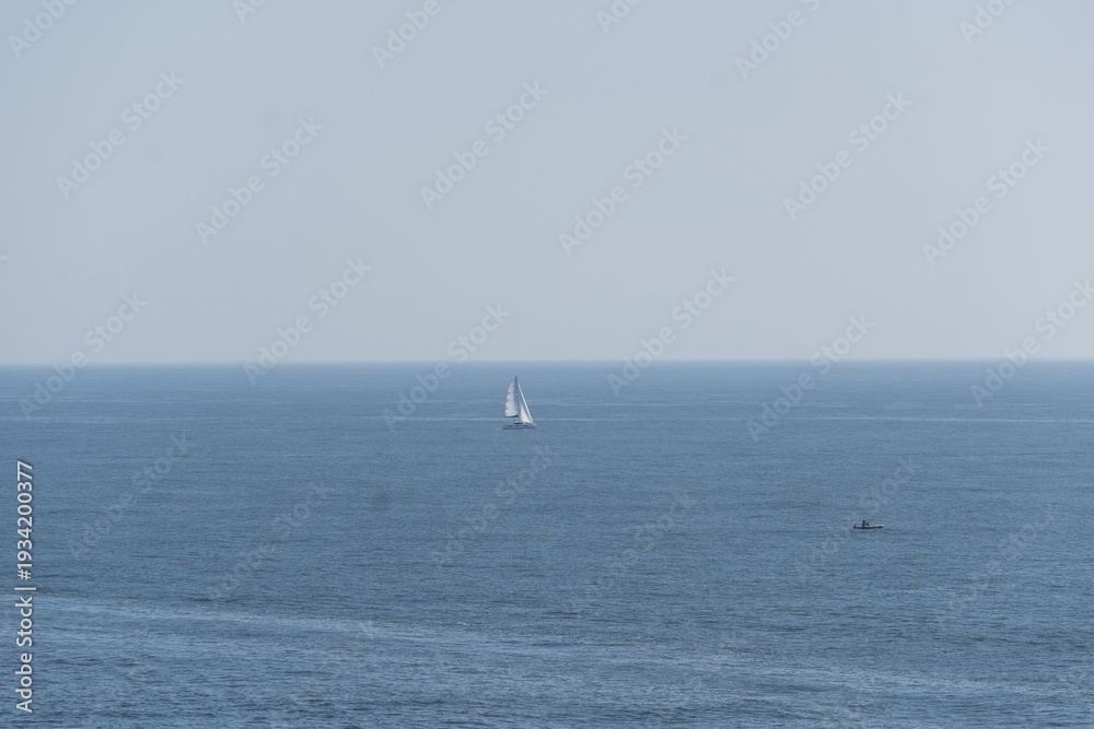 Fototapeta premium Sailboat gliding across the calm ocean waters under a clear blue sky. Yacht with white sails vast expanse of blue water. Background with faint outline of distant land, creating a serene maritime scene