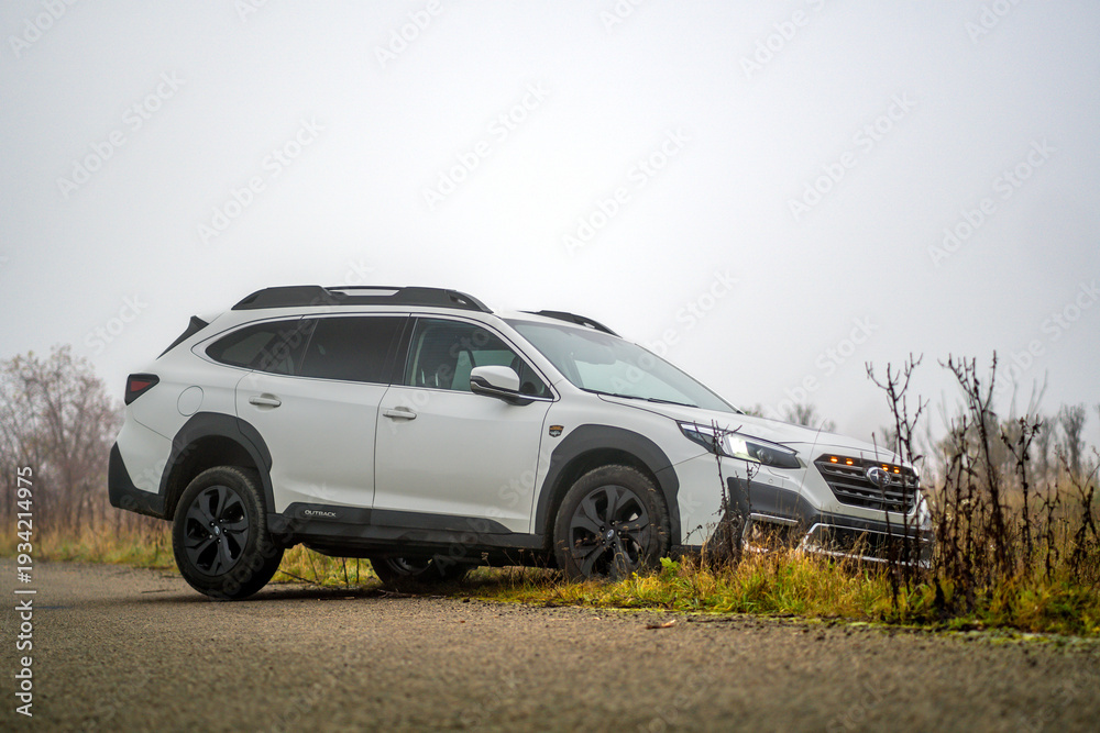 Fototapeta premium Brno, Czech Republic - November 12, 2025: White Subaru Outback family car driving from field to road in autumn in fog outdoors. Japanese off-road SUV.