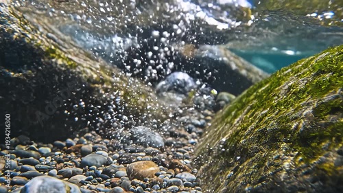 Underwater view of a clear river stream with smooth pebbles on the riverbed and abundant air bubbles rising to the surface through mossy rocks.