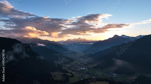 Majestic mountain valley bathed in the warm glow of sunrise with dramatic clouds and mist swirling around the peaks creating an ethereal and aweinspiring landscape.