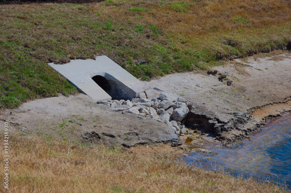 Fototapeta premium Concrete Culvert Drain at Rocky Shoreline Retention Pond With Erosion and Runoff Near Dry Grassy Bank rock riprap on a dry grassy bank beside a shallow shoreline, showing erosion, stormwater runoff a