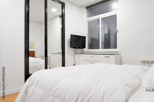 Hotel room with white bedding, wall mounted television and mirrored wardrobe. Simple affordable accommodation interior with window
