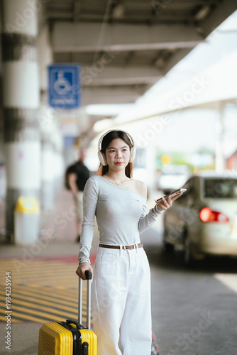Young Asian woman with headphones holding smartphone and suitcase at airport pickup area, waiting for transport with cars in background.