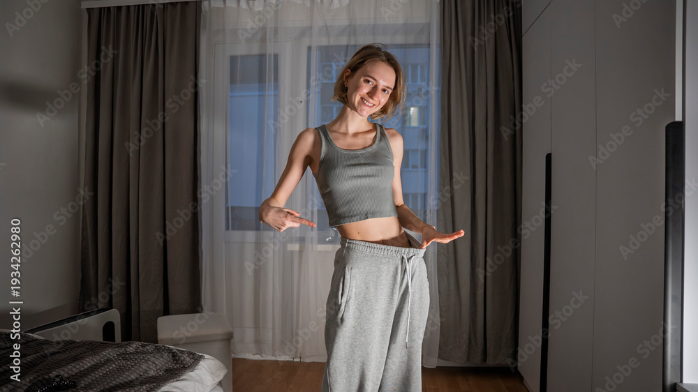 Fototapeta premium Happy young woman pointing at her loose gray sweatpants, showing significant weight loss results. Concept of successful dieting, fitness progress, and body transformation before spring season.
