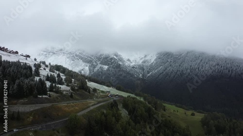 A scenic view of the French Alps featuring snow dusted mountains, evergreen forests, and a green valley under cloudy skies. Early winter atmosphere in a tranquil alpine setting.
