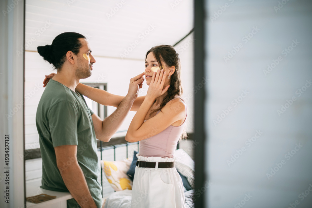 Fototapeta premium Family spa day at home. Young woman and man with golden hyaluronic patches on their faces in the bedroom of their home.