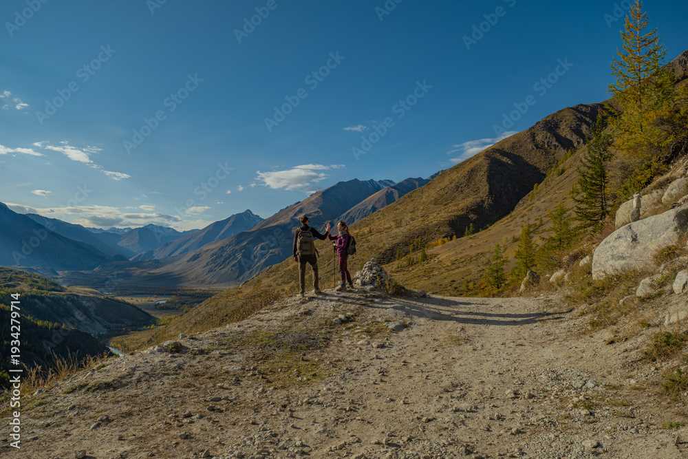Fototapeta premium Couple of hikers with backpacks giving high five on mountain edge overlooking scenic alpine valley during autumn trekking trip for teamwork, success and travel adventure concept.