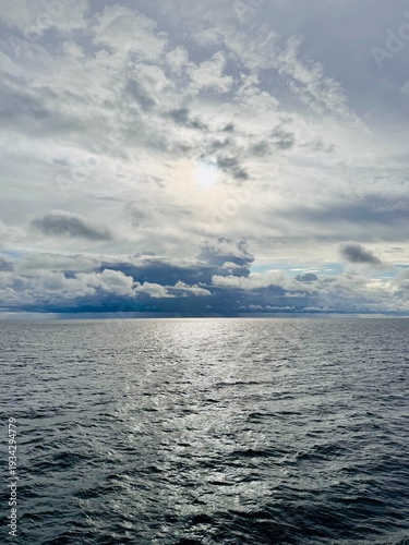Wide view of open sea under a dramatic overcast sky with layered clouds and diffused sunlight breaking through. The light reflects softly on the textured water surface, creating a calm yet atmospheric