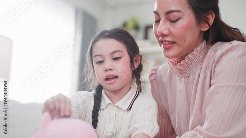 Closeup Asian mother teaches her young daughter how to save money by putting a coin into a pink piggy bank together in a bright modern living room.