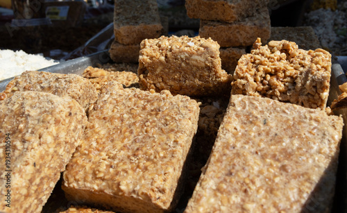 High-angle close-up of traditional Brazilian paçoca peanut candy blocks. Rustic sweets, made from peanuts and sugar, show brown texture. Bright sunlight creates strong shadows on a market tray.