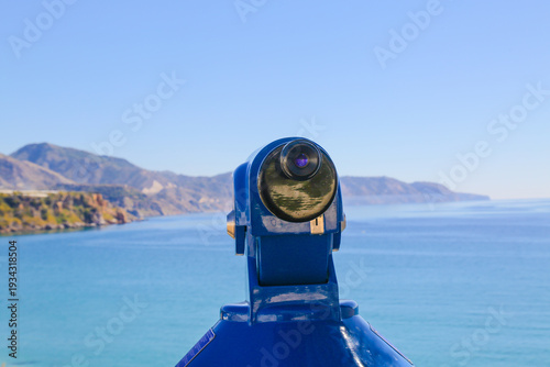 Blue coin-operated telescope on a scenic coastline overlooking the ocean and mountains