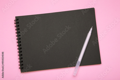 Black notebook with black pen on pink background. Mockup