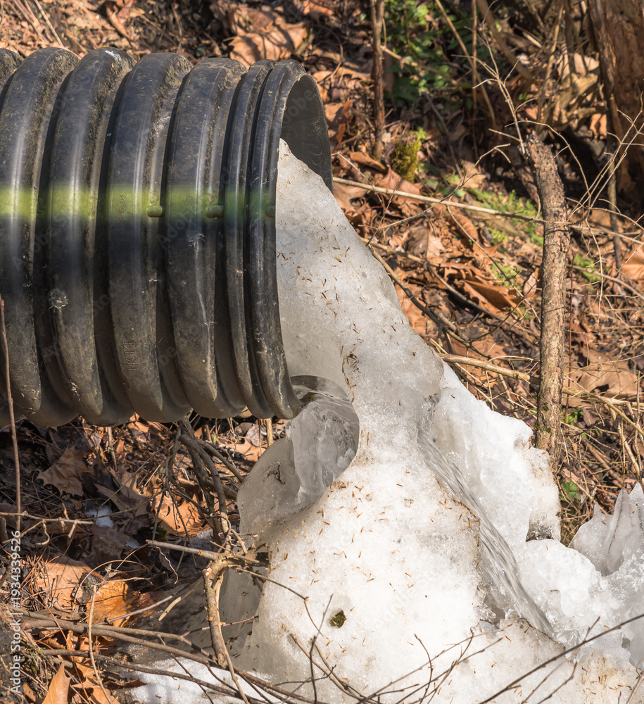 Obraz premium A corrugated drainage pipe discharging meltwater and ice into a small creek in in Warren County, Pennsylvania, USA on a sunny winter day