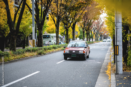 an old red car driving down a tree-lined street on a bright day. The image captures the essence of classic cars and the beauty of an autumn landscape in Kyoto, Japan