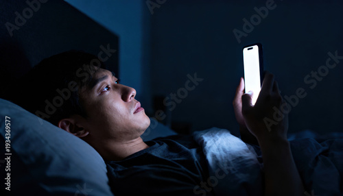 Man lying in bed at night scrolling smartphone with bright screen lighting face, sleepless and focused