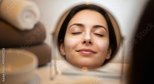 Beautiful young woman looking in mirror with eyes closed and face relaxed, ready for skincare treatment with cream on her face