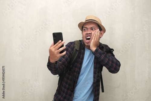 Southeast Asian man in straw hat and backpack reacts angrily to a smartphone, holding his cheek in shock over bad travel news.