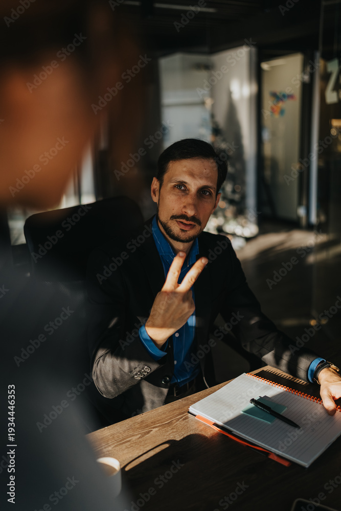 Fototapeta premium A man in a dark suit sits at a wooden table with a notebook and pen. He shows a peace sign with two fingers in a casual office setting.
