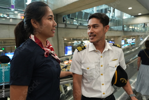 An Asian female flight attendant and a male captain walk and chat up the escalator; a service team in the airline industry.
