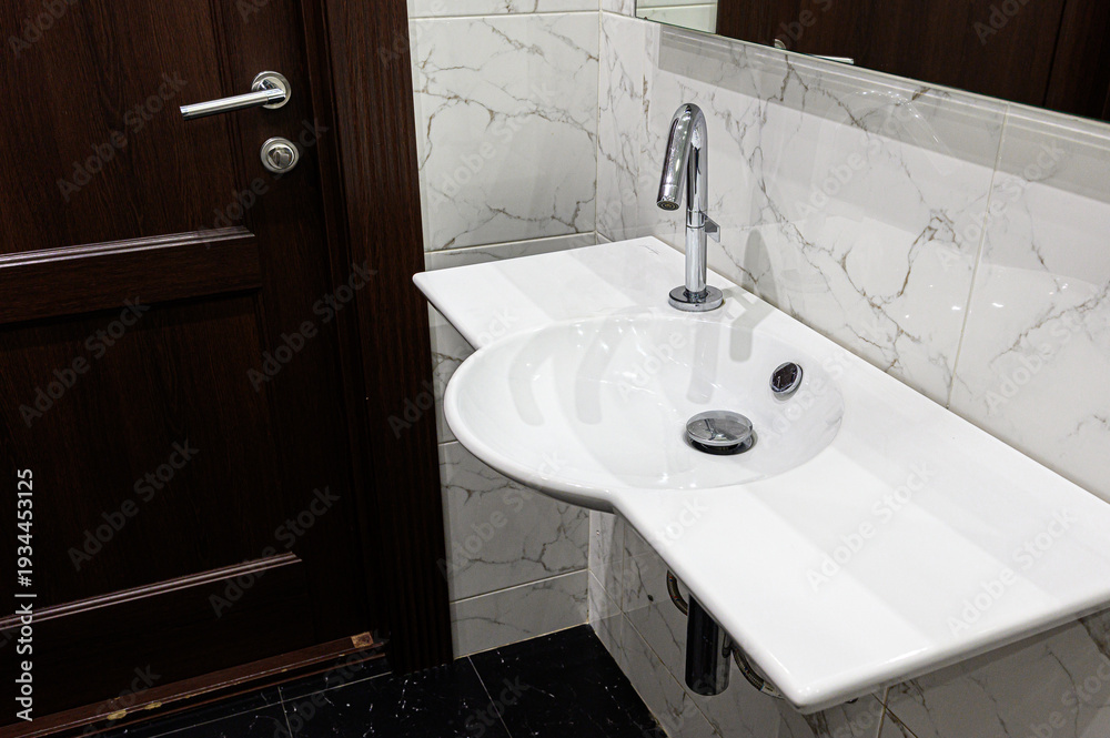 Fototapeta premium A modern bathroom sink with a sleek chrome faucet mounted on a white ceramic countertop. Set against marble-patterned tiles, next to a dark wooden door with a metallic handle.