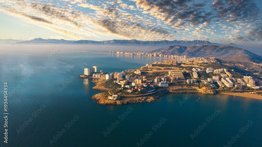 Fototapeta premium Aerial drone view of Cullera Faro district on the Mediterranean coast of Spain. Golden hour light illuminates the peninsula, coastal apartments and mountains with calm sea and misty horizon.