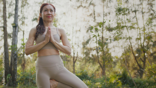 Woman in beige activewear standing in tree pose with hands in prayer at sunrise in forest, calm and focused expression