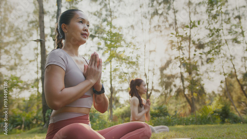Two women meditate in outdoor park at sunrise with hands pressed together, calm focus, soft light and yoga mat on grass