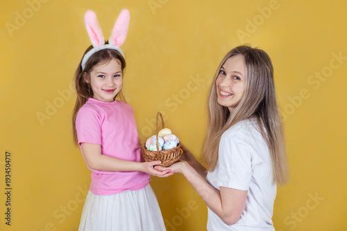 Woman and girl with bunny ears present a basket filled with colorful Easter eggs, set against a vibrant yellow backdrop, highlighting a joyful holiday spirit