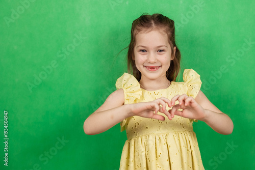 Little girl with brown hair wearing a yellow dress makes a heart shape with her hands against a green background in a studio setting