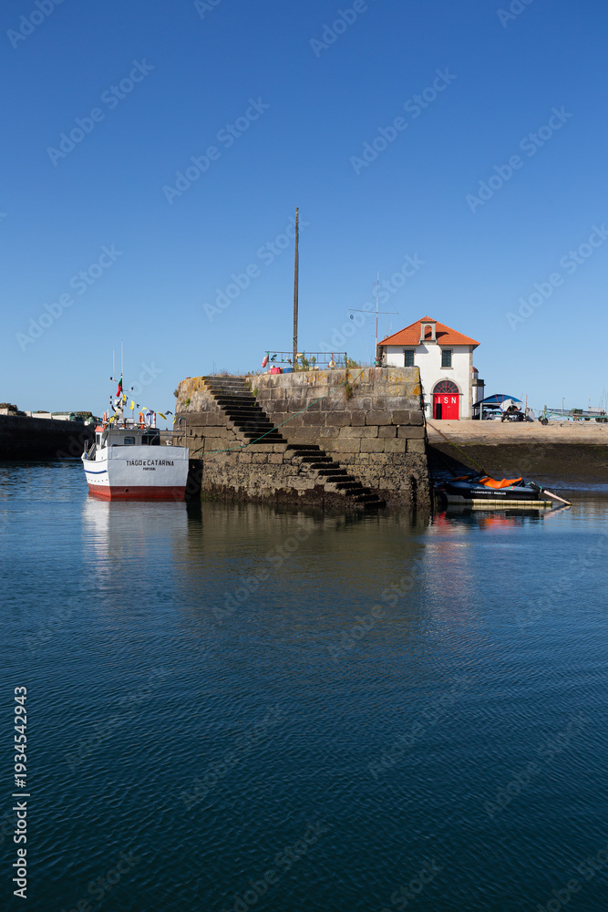Fototapeta premium 20 August 2024: The ISN station at the fishing quay in Viana do Castelo stands prominently by the waterfront, its red doors and white facade reflecting in the calm blue harbor waters, Viana do Castelo