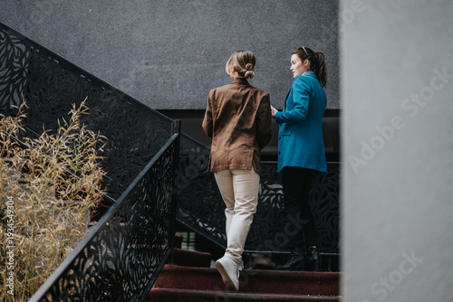 Two women converse on a decorative stair rail in an urban setting. Two women talking on stairs in a modern, outdoor space.
