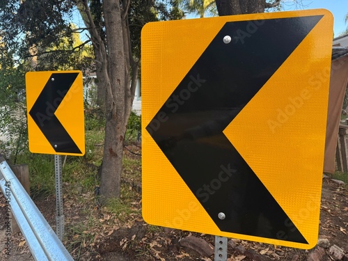 Принти на полотні Sharp Curve Warning Signs: Large yellow chevron road signs indicate a dangerous left-hand curve ahead, protecting residential yards from oncoming traffic at a junction