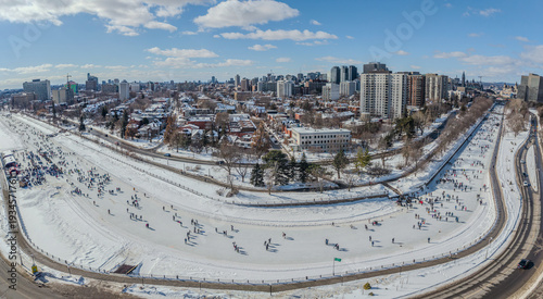 Panoramic drone view of skaters on the frozen Rideau Canal Skateway in downtown Ottawa, Ontario, Canada, capturing winter recreation, urban skyline, snowy landscape, vibrant city life (February 2026).