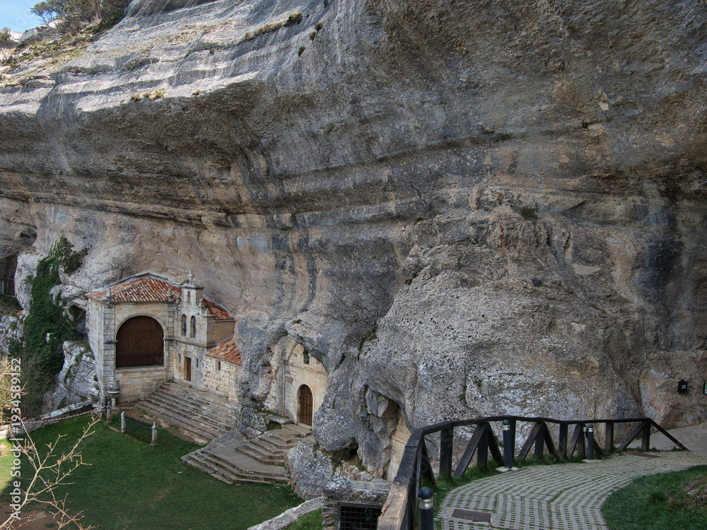 Obraz premium San Bernabé Hermitage in Ojo Guareña - Historic Hermitage of San Bernabé built into the massive limestone rock face and cave entrance of the Ojo Guareña complex in Burgos, Spain.