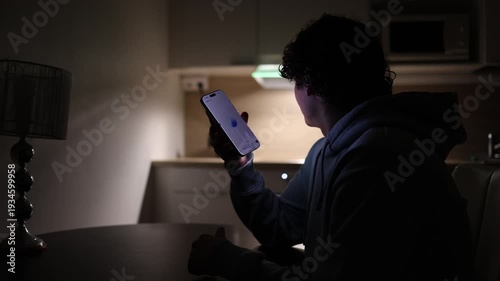 Young student at desk in dark apartment studying at night using smartphone communicating with chatgpt asking questions ai assistant helping with education and homework