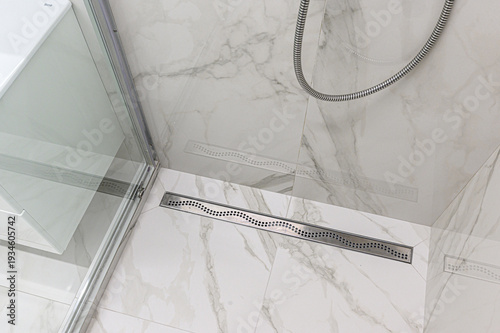 Obraz na płótnie A modern shower area with marble-patterned tiles, linear stainless steel drain, glass door, and flexible chrome shower hose mounted on the wall