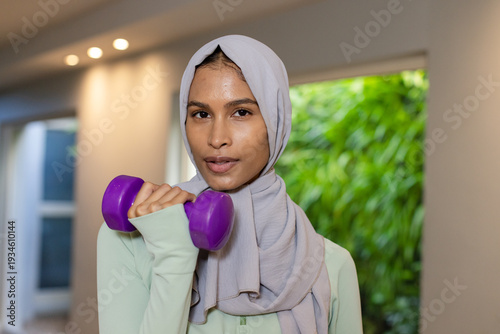 Adult female wearing light-gray hijab and pale-green top lifting purple dumbbell in home studio