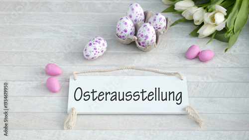 Heller Hintergrund mit Ostereiern und Blumen auf einem Schild steht der Schriftzug Oster-Ausstellung.