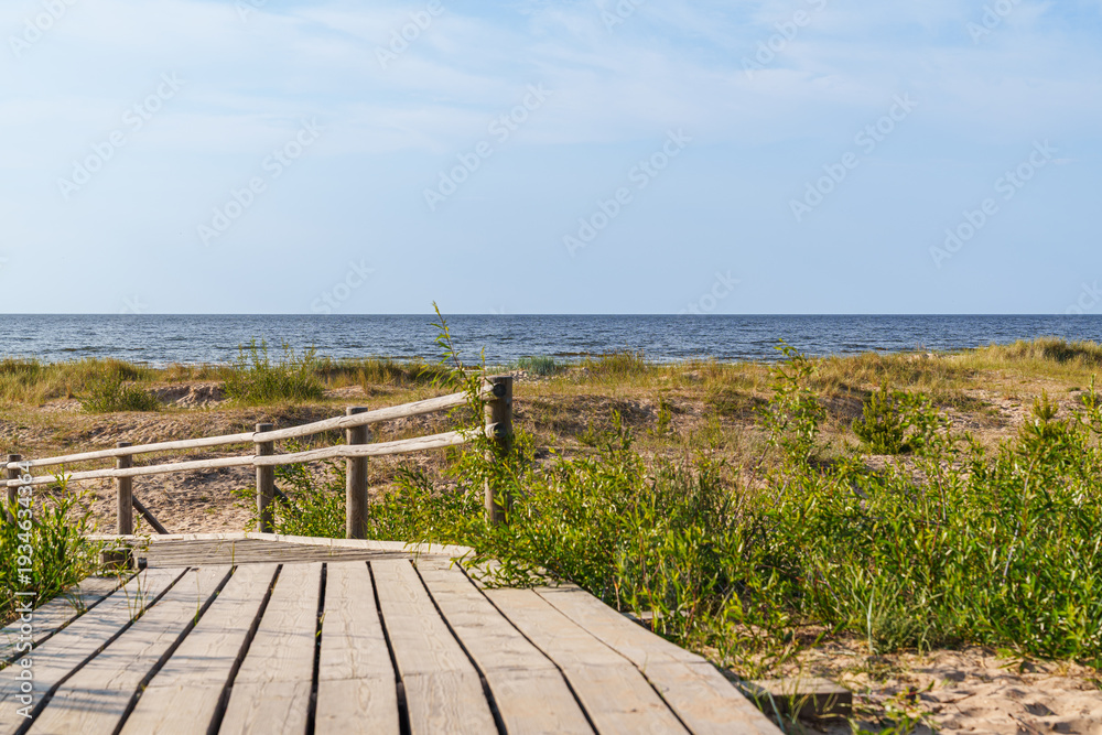 Fototapeta premium A wooden boardwalk leads to the Baltic sea in Latvia. The beach is covered in grass and there is a clear blue sky above