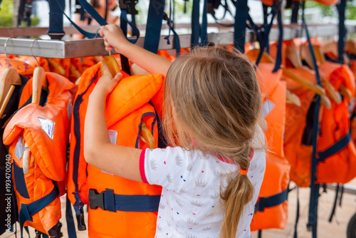 Obraz na plátně little girl choosing life jacket for boating, orange life vests hanging, Safety