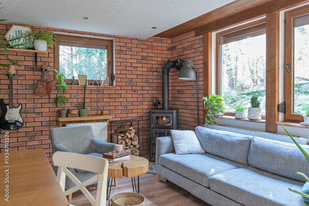 Fototapeta premium Rural cottage with cozy design, windows to the forest, fireplace, sofa, armchair and red brick wall. Interior of living room with design indoor architecture.