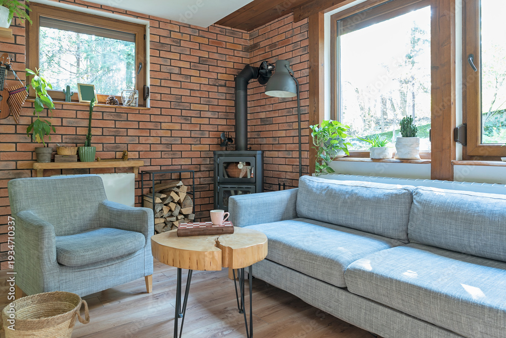 Fototapeta premium Rural cottage with cozy design, windows to the forest, fireplace, sofa, armchair and red brick wall. Interior of living room with design indoor architecture. 
