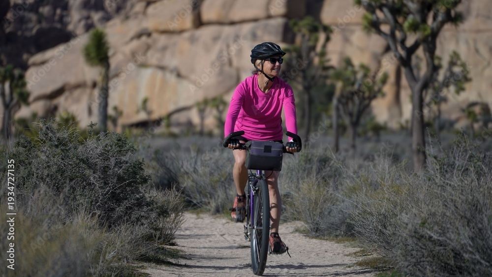 Fototapeta premium A mature woman rides her bike on a dirt road in Joshua Tree National Park, surrounded by unique rock formations and plants