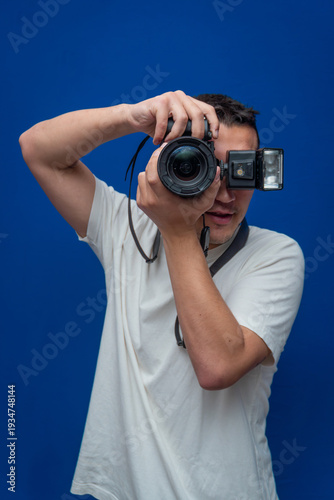 Male photographer taking a picture, holding a professional digital camera with an attached flash, standing against a blue background