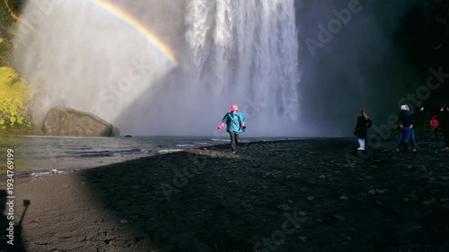 Running happy female tourist on visit of the Skogafoss waterfall flowing from cliff in Iceland, a famous place and travel destination, slow motion